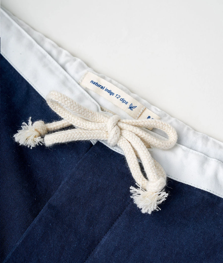 A white cotton garment with blue "Natural Indigo" tag, fastened with a braided drawstring against a navy blue background - Industry of All Nations