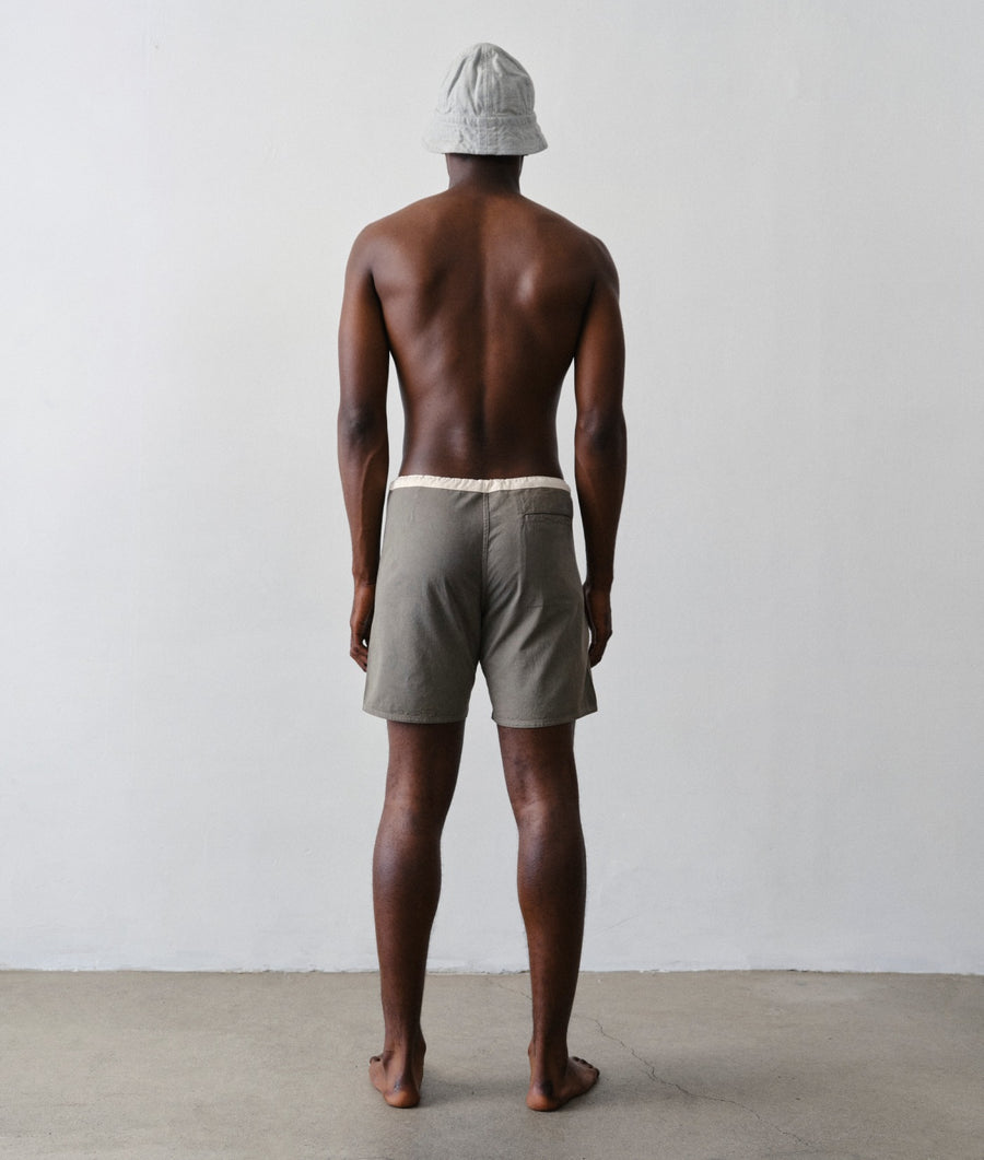 Back view of a shirtless man wearing IOAN Contrast Boardshorts in Iron 4 and a light gray bucket hat, standing against a plain white wall.
