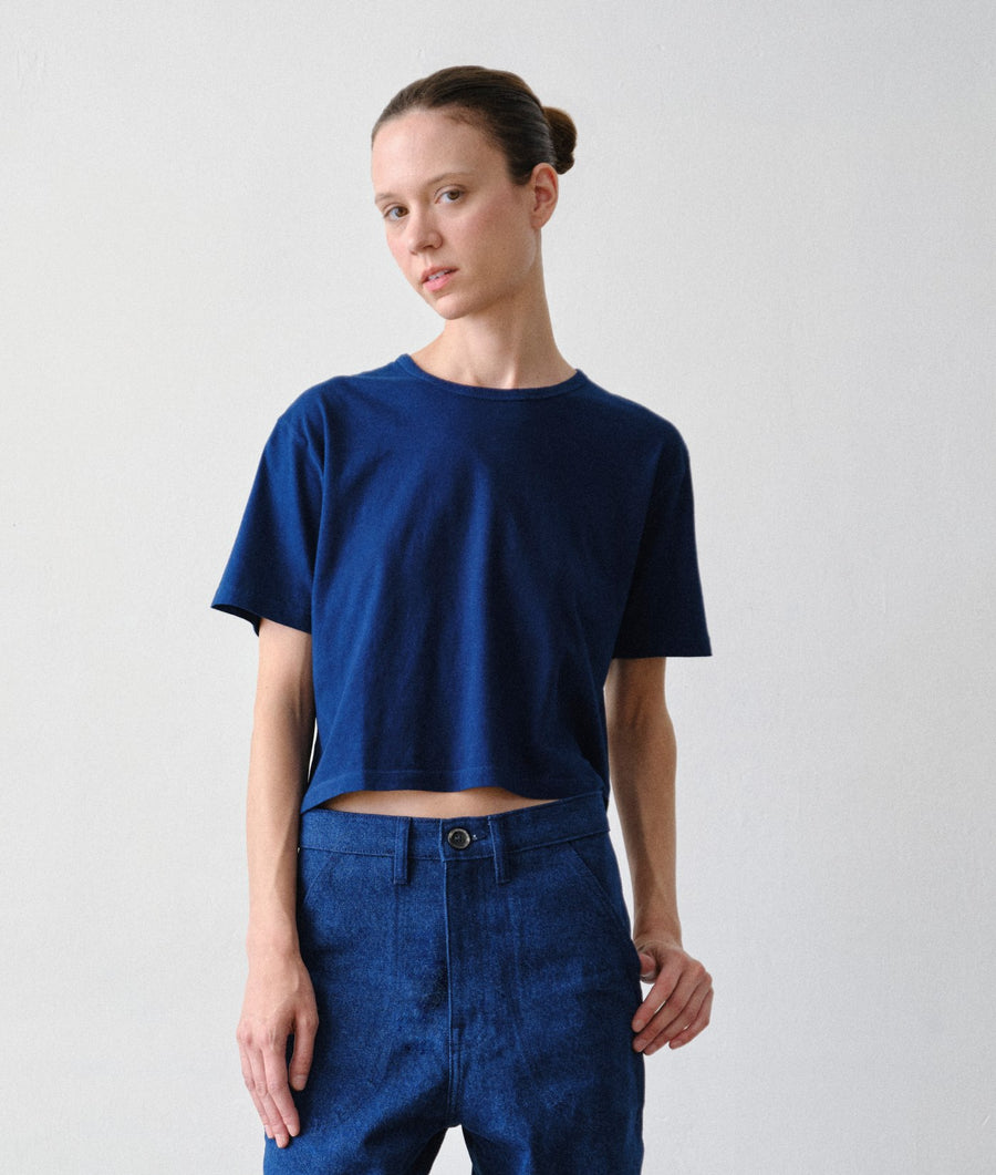 A woman wearing the IOAN Cropped Short-Sleeve T-Shirt in Indigo 12 and matching IOAN Upcycled Blue Jeans stands against a plain light gray studio background.
