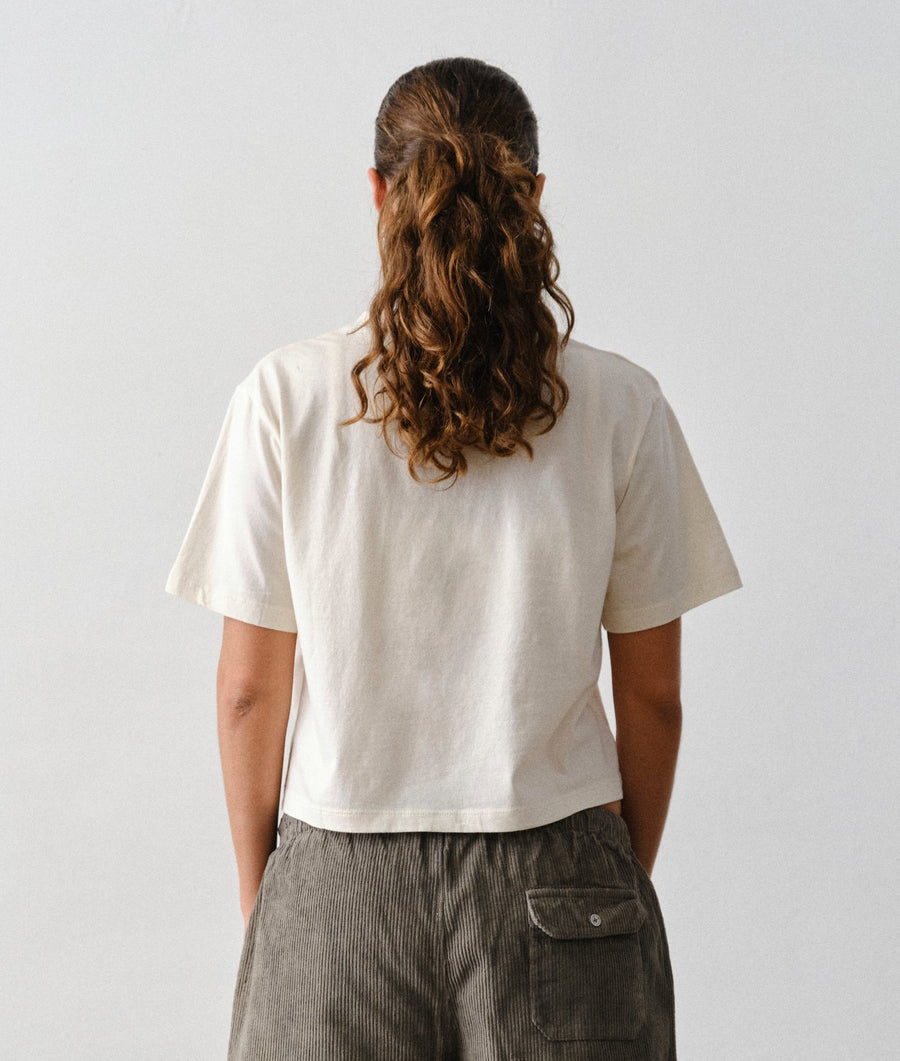 A woman wearing an IOAN undyed cropped T-shirt and corduroy pants, viewed from the back against a plain light gray background.