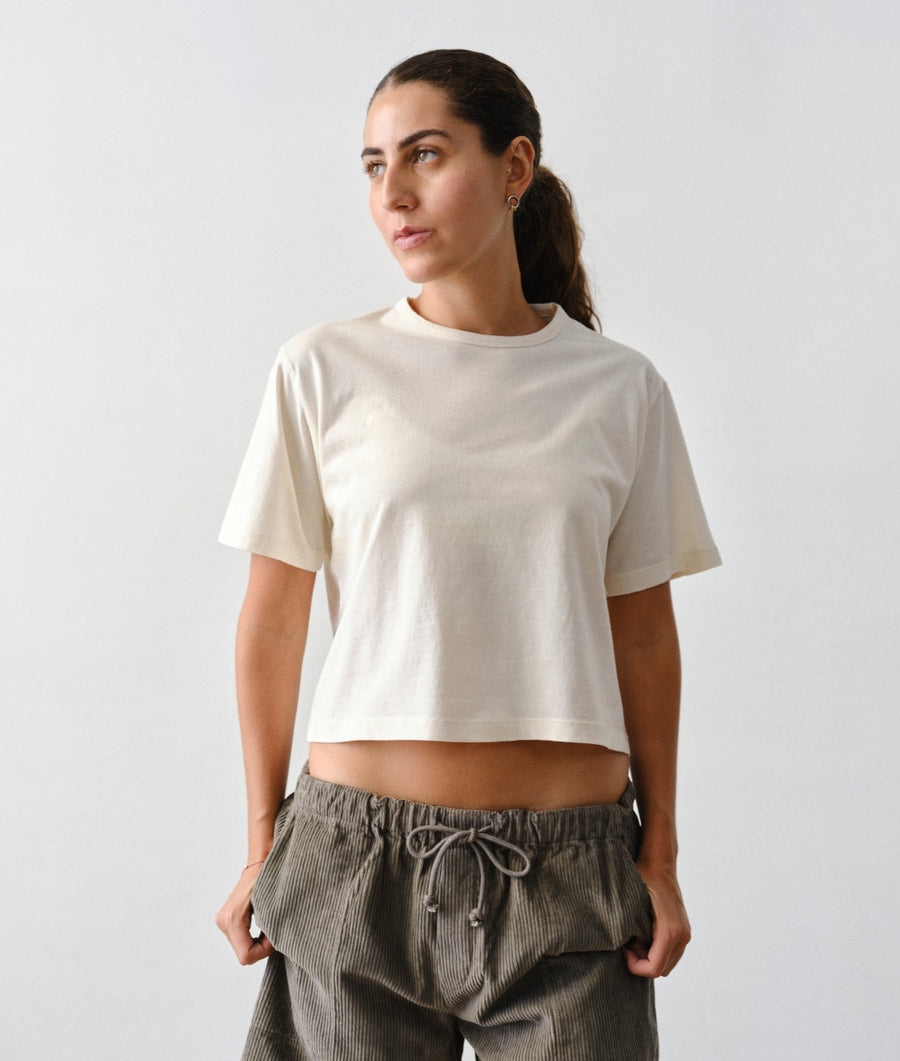 A woman wearing an IOAN undyed cropped T-shirt and gray corduroy drawstring pants against a light gray studio background.