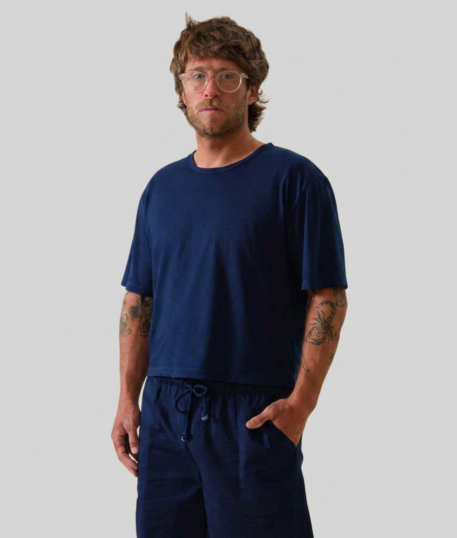 A man wearing an IOAN Indigo 12 cropped T-shirt and drawstring pants, standing against a solid light gray background.