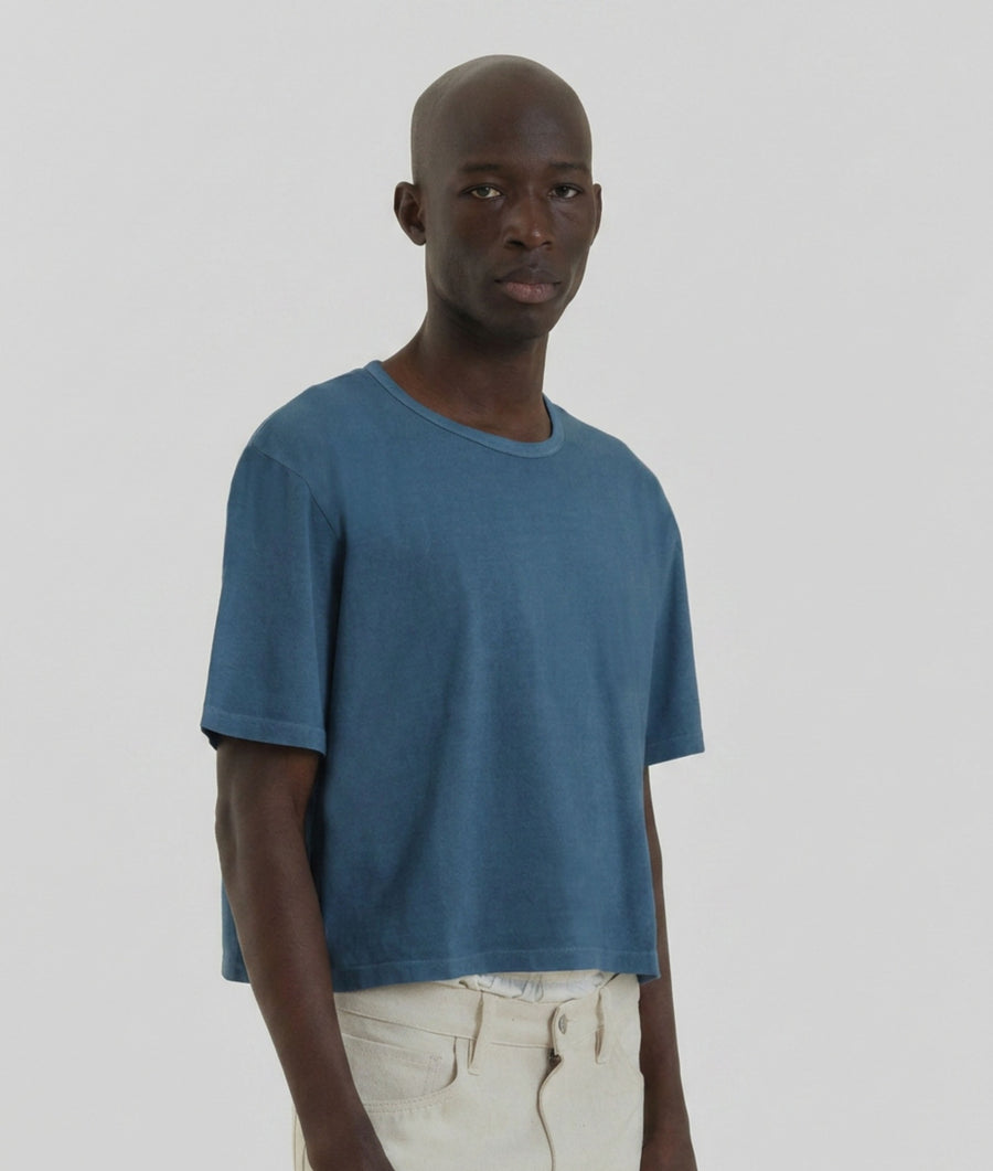 A man wearing an IOAN Indigo 6 cropped T-shirt and undyed pants, standing against a solid light gray background.