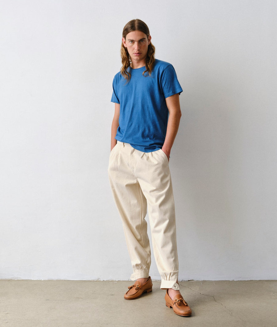 A model standing against a plain light gray wall wearing an IOAN Indigo 6 short sleeve crewneck T-shirt, Gaucho Pants in undyed cotton, and brown leather loafers.