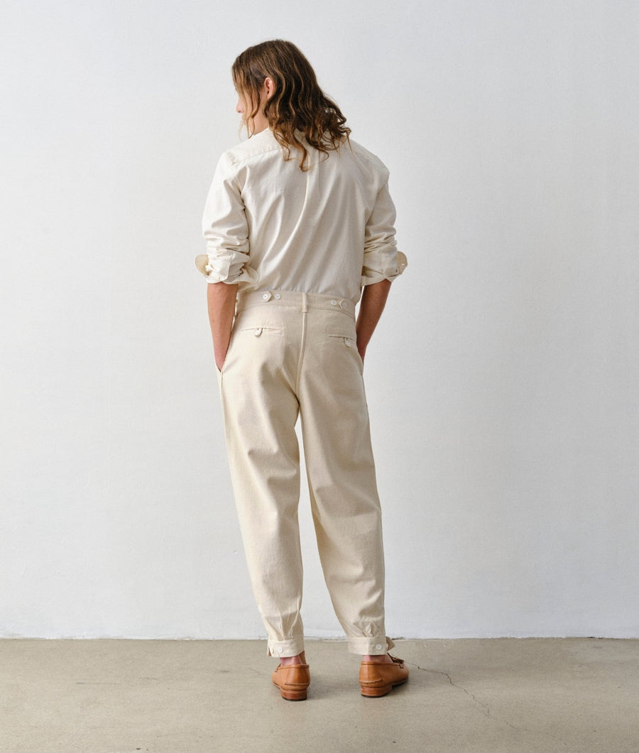 A man stands with his back to the camera against a plain white wall wearing undyed IOAN Gaucho Pants, an undyed IOAN Madras Shirt, a gray belt, and brown leather loafers.