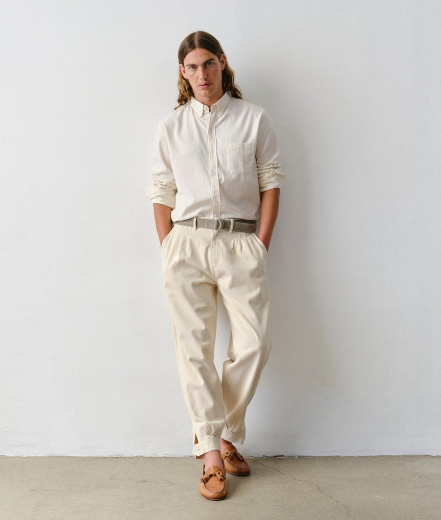 A man stands against a plain white wall wearing undyed IOAN Gaucho Pants, an undyed IOAN Madras Shirt, a gray belt, and brown leather loafers.