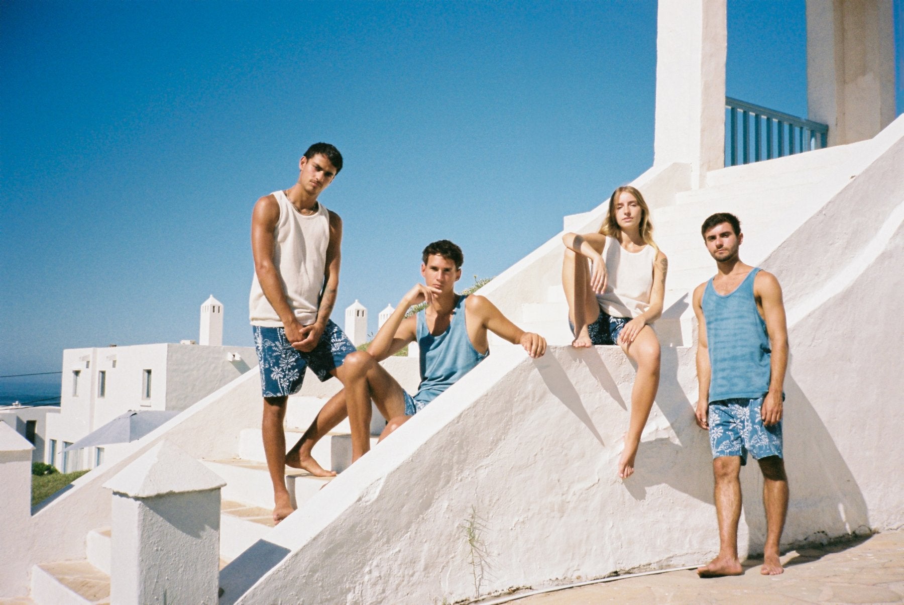 Four young men wearing swim trunks and tankinis posing on white outdoor stone steps under a clear blue sky.