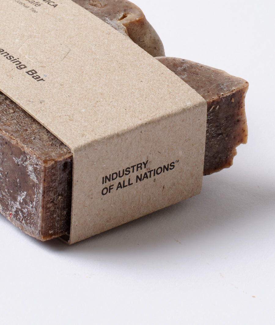 Product packaging featuring the text "Industry of All Nations" on a brown cardboard material, with a piece of handcrafted, natural-colored fabric visible in the background. - Industry of All Nations