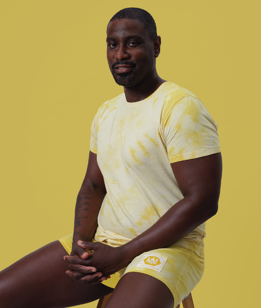 A middle-aged Black man wearing a yellow organic cotton t-shirt with a patch logo, sitting against a yellow background - Industry of All Nations