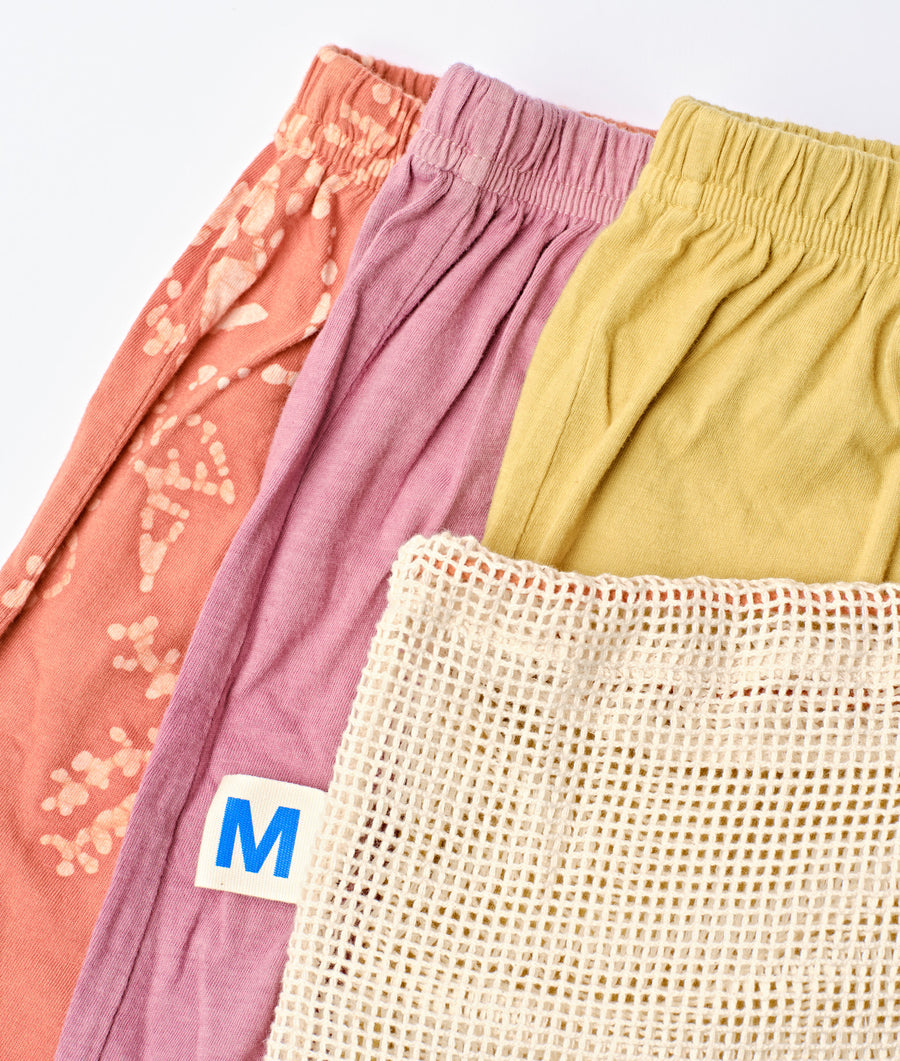 Flat lay of three brightly colored organic cotton skirts, along with a woven mesh laundry bag with an 'M' label - Industry of All Nations