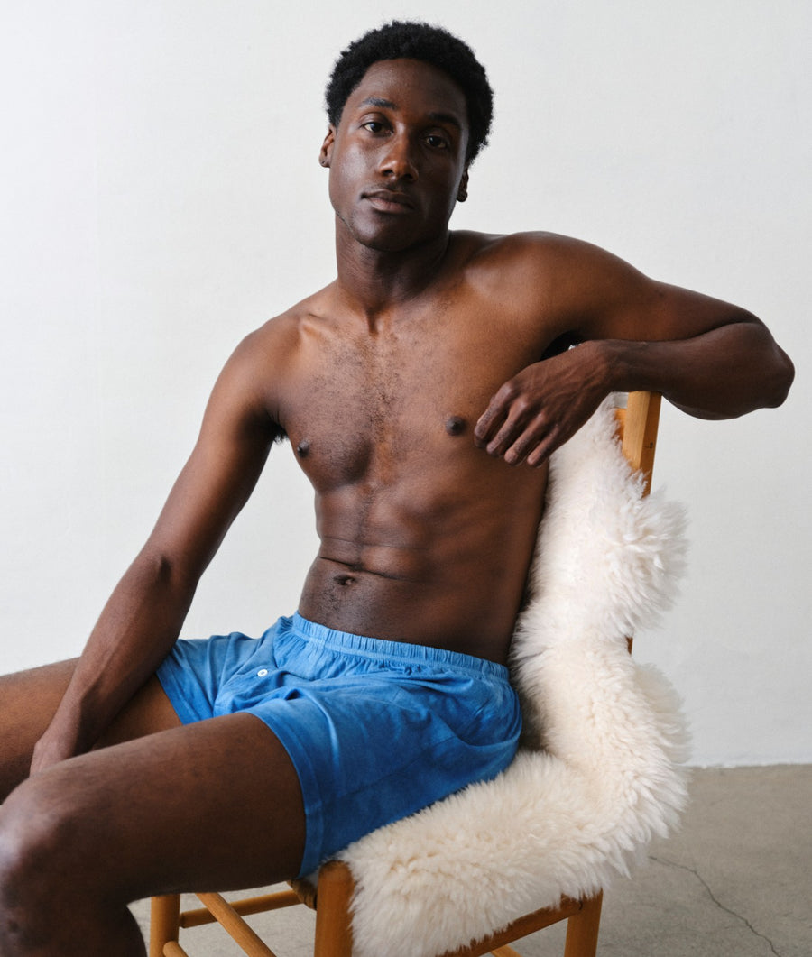 A shirtless man sitting on a wooden chair with a white faux-fur cushion, wearing bright blue swim trunks against a plain light gray wall.