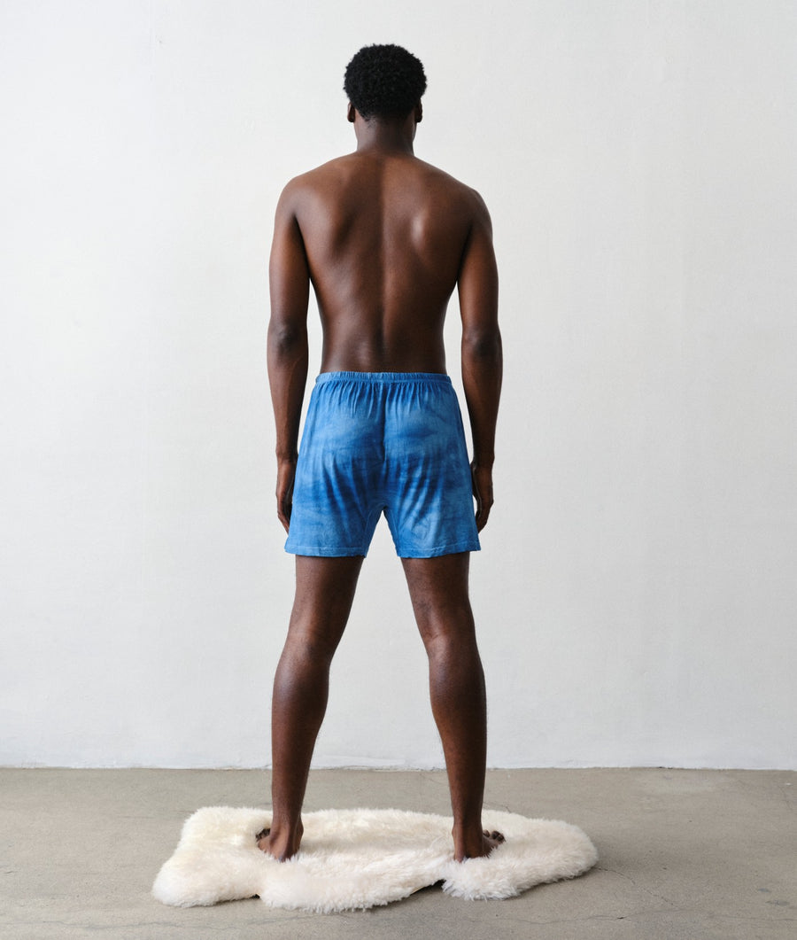 Man standing with his back to the camera wearing IOAN Indigo 6 boxer briefs on a white faux-fur rug in a minimalist studio with a light gray wall and concrete floor.