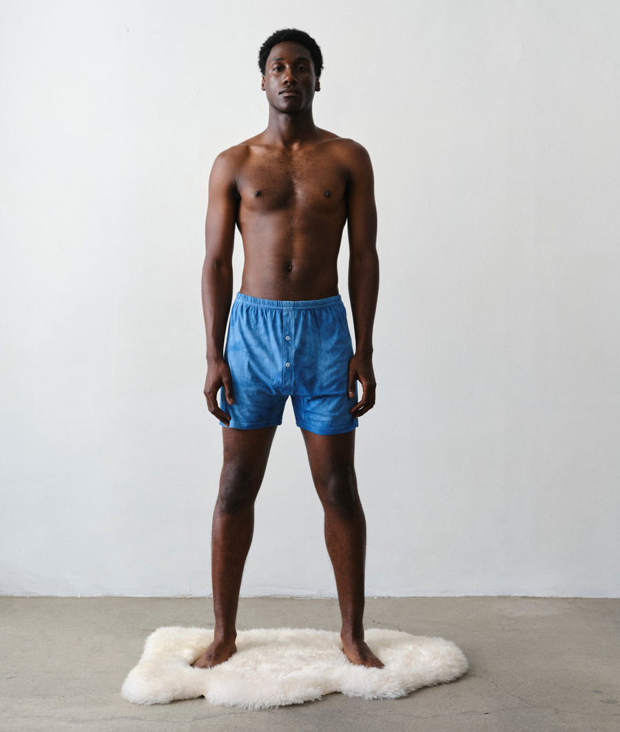 Man standing facing the camera wearing IOAN Indigo 6 boxer briefs on a white faux-fur rug against a light gray wall with a concrete floor.