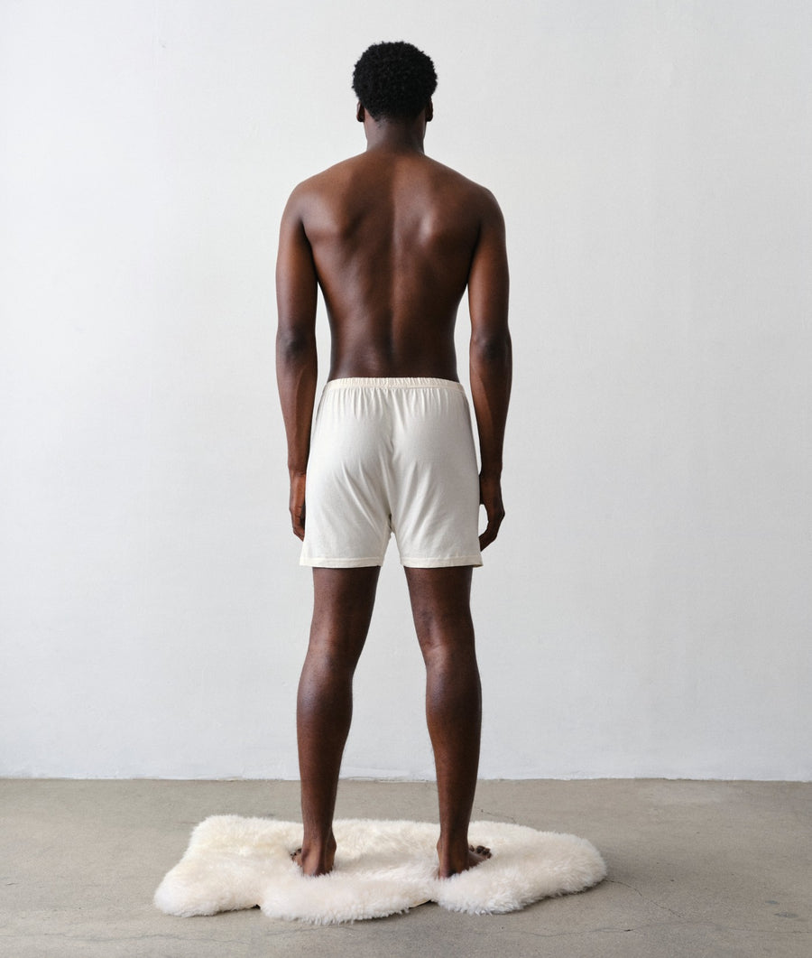Man standing with his back to the camera wearing IOAN undyed boxer briefs on a white faux-fur rug against a light gray wall with a concrete floor.