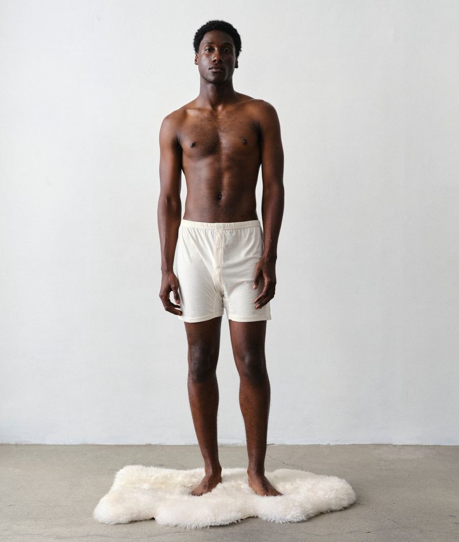 Man standing facing the camera wearing IOAN undyed boxer briefs on a white faux-fur rug against a light gray wall with a concrete floor.