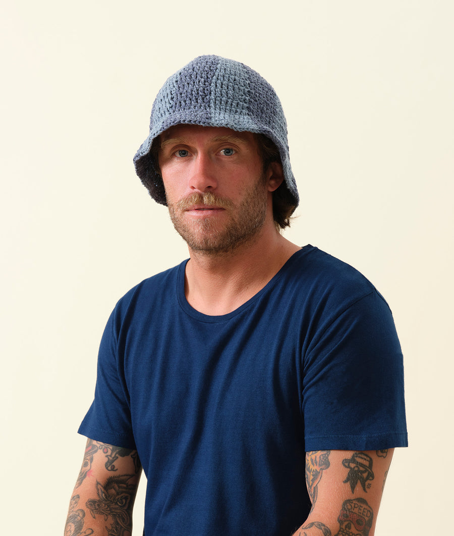 A man wearing a gray crocheted bucket hat, with a blue t-shirt and tattoos on his arms - Industry of All Nations