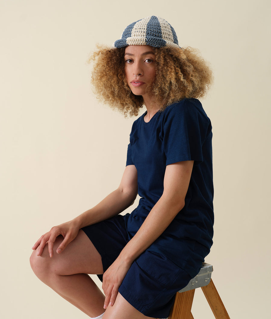A young woman wearing a navy blue dress and a crocheted patterned hat, posing on a stool - Industry of All Nations