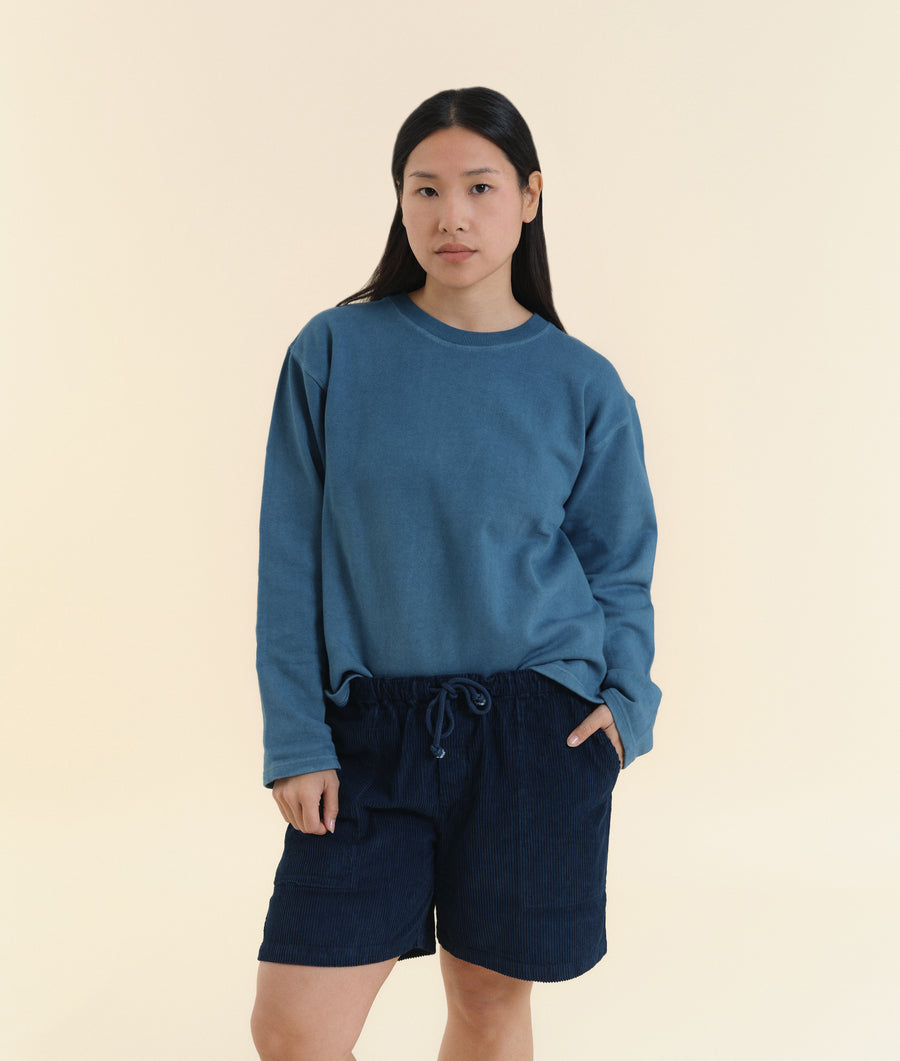 A young woman wearing a blue cotton sweatshirt and navy blue shorts, standing against a light beige background - Industry of All Nations