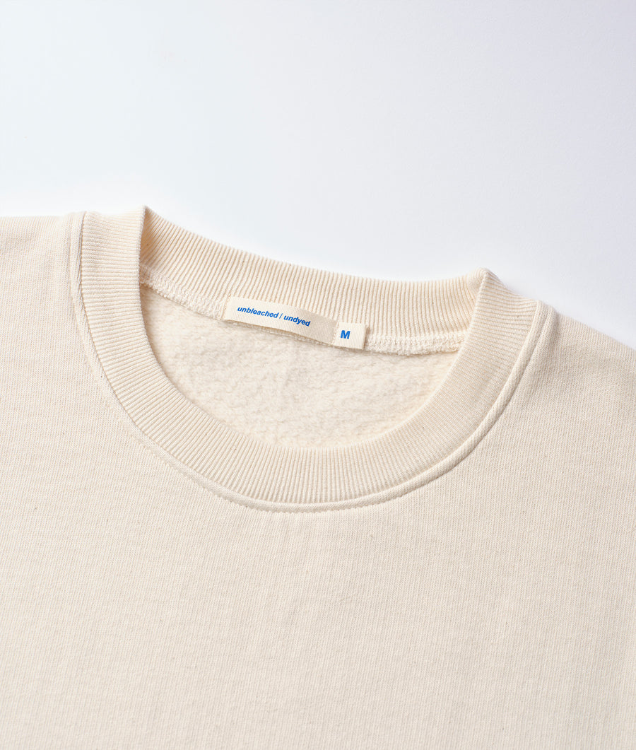 Cream-colored crewneck t-shirt with a tag that reads "unbleached/undyed" displayed in a flat lay shot - Industry of All Nations