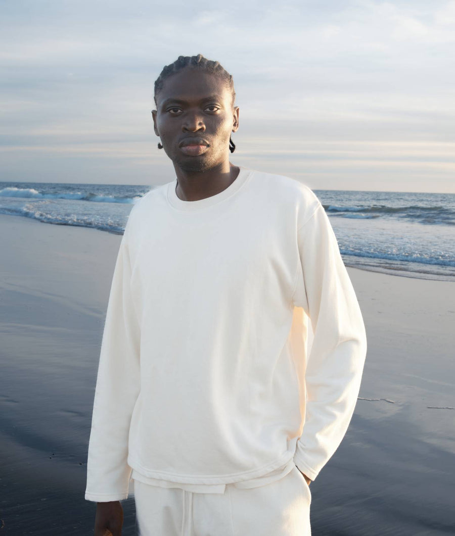 A young man wearing a white long-sleeve top standing on a beach overlooking the ocean - Industry of All Nations