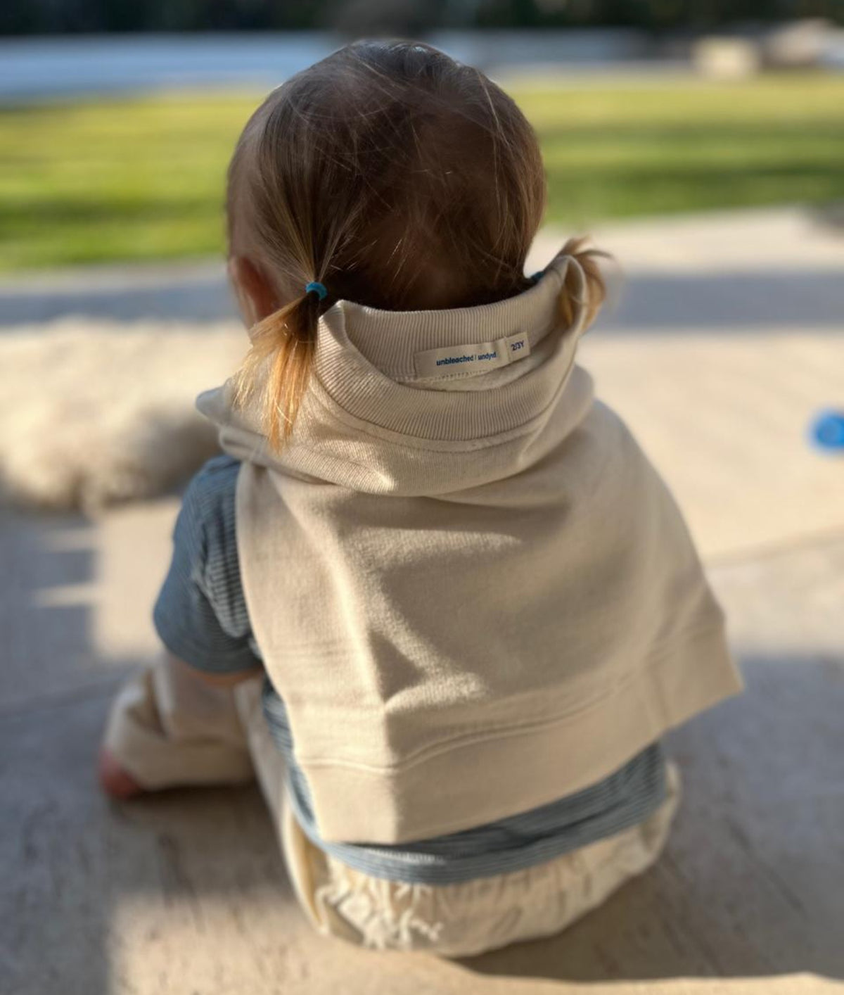 A toddler sitting outdoors on a sunny day, viewed from behind. The child is wearing a light beige hooded sweatshirt with a built-in neck warmer, layered over a blue knit top and cream pants.