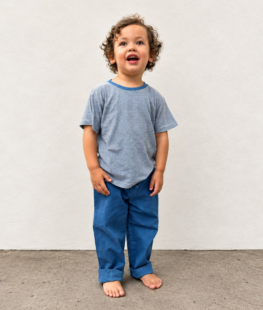 A toddler standing barefoot against a light gray wall, wearing a short-sleeve heathered blue-gray t-shirt with a blue collar and blue pants.