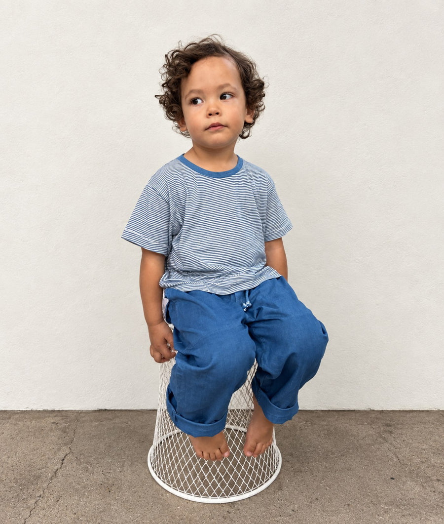 A toddler with curly brown hair sits barefoot on a white wire stool, wearing a light blue and white striped short-sleeve t-shirt and blue pants, set against a plain off-white wall and concrete floor.