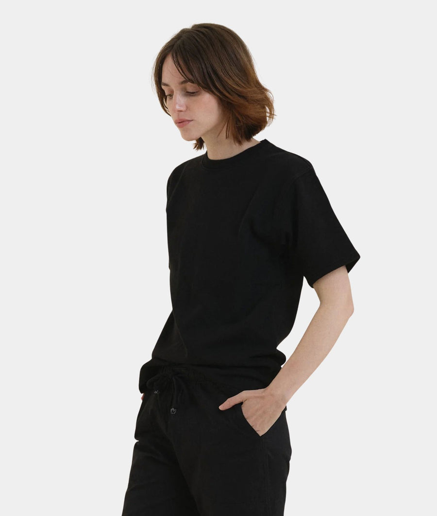 A woman with short brown hair wearing IOAN Iron 8 Kung Fu T-Shirt and black pants, standing against a light gray studio background.