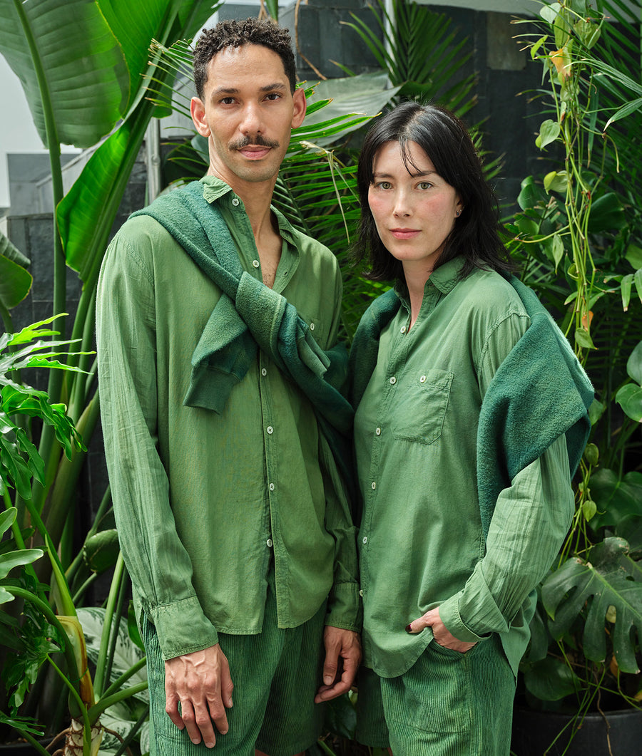 A man and woman wearing matching green organic cotton button-down shirts in a lush green environment - Industry of All Nations