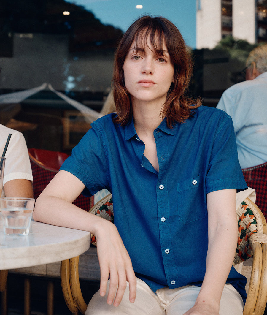 The image shows a woman wearing a blue short-sleeved button-down shirt from Industry of All Nations.
