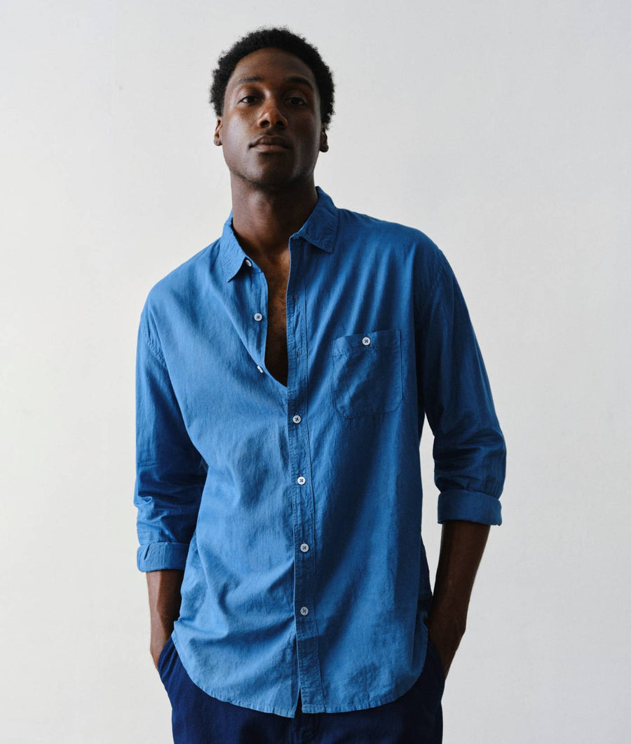 A man wearing a medium blue long-sleeve button-down shirt with a chest pocket, standing against a plain light gray background.
