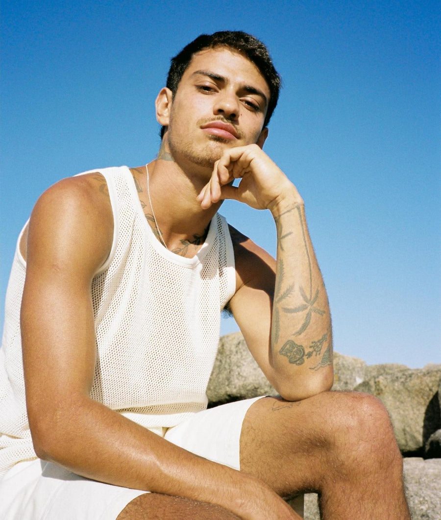 A man wearing a white mesh tank top, sitting with a pensive expression against a blue sky background - Industry of All Nations