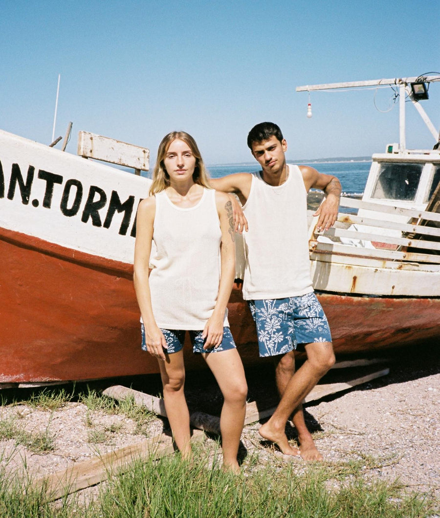 A man and woman standing next to a boat, wearing white tank tops and patterned shorts in a coastal setting - Industry of All Nations