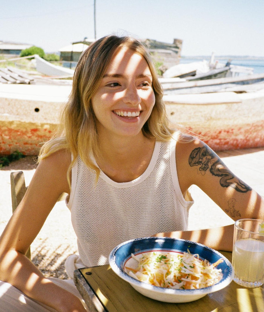A smiling woman wearing a white mesh tank top, sitting outdoors with a bowl of food - Industry of All Nations