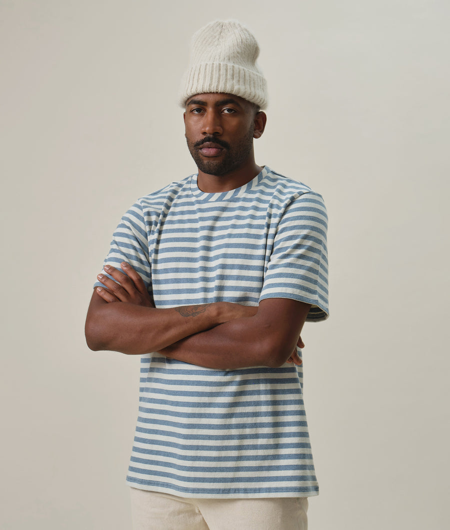 A man wearing a striped blue and white t-shirt and a white knit hat, standing with arms crossed - Industry of All Nations