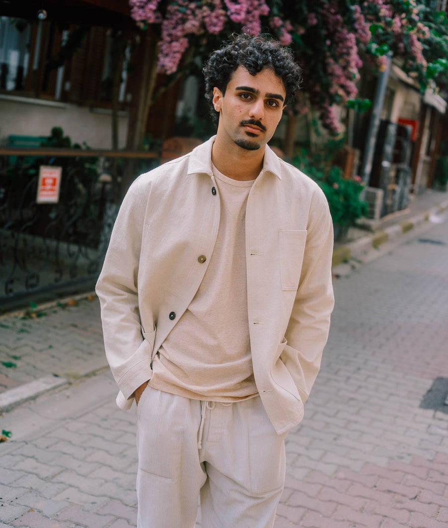 A person wearing a cream-colored button-up coat and pink sweater, standing outdoors in front of pink flowers - Industry of All Nations