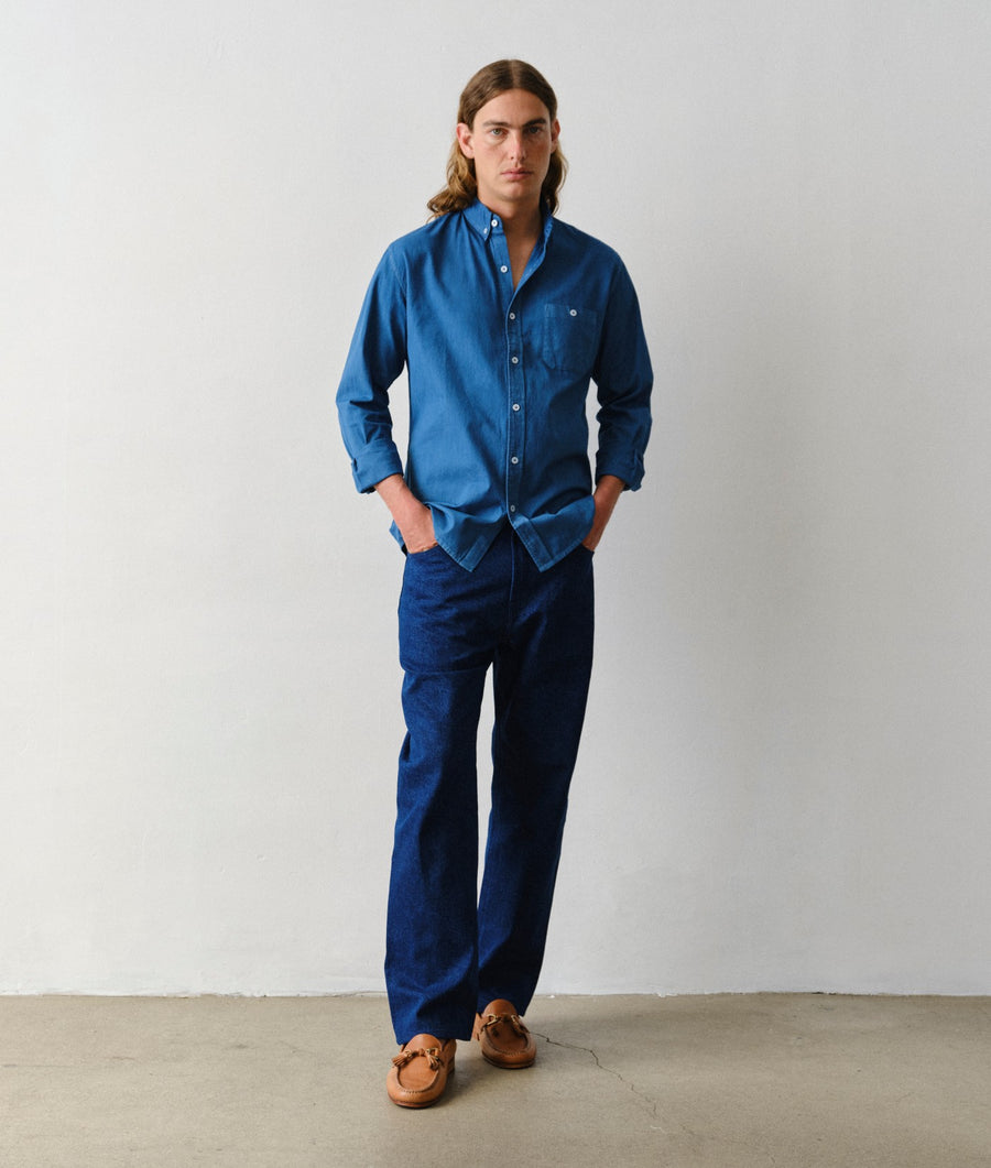 Man wearing an IOAN Madras Long-Sleeve Button-Up Shirt in Indigo 12 and matching New Jeans in Azul, standing with his hands in his pockets against a plain light gray wall.