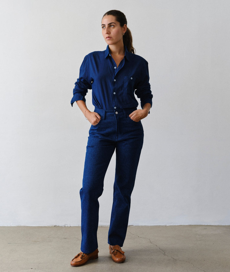 A woman modeling IOAN New Jeans in Azul with an IOAN Madras Shirt in Indigo 12 and brown leather shoes, standing against a plain white wall.