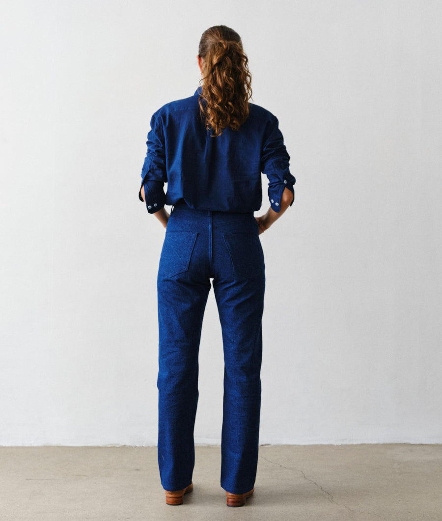 Rear view of a woman modeling IOAN New Jeans in Azul with an IOAN Madras Shirt in Indigo 12 and brown leather shoes, standing against a plain white wall.