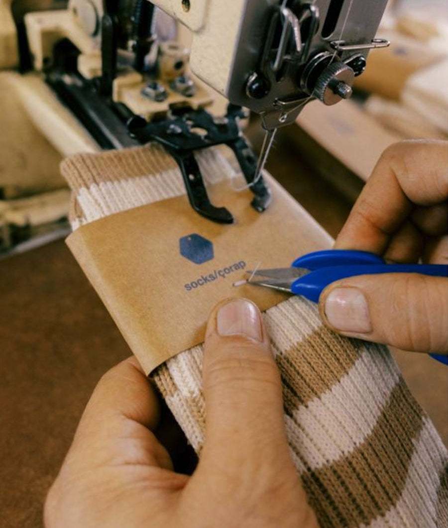 Close-up of a maker in Turkey preparing IOAN Organic Cotton Socks, guiding a brown paper sleeve over the beige and brown striped knit socks, with a sewing machine and scissors visible in the background.