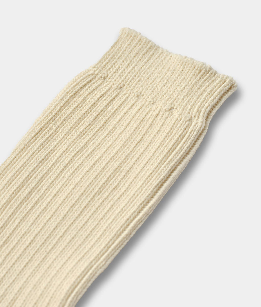 A close-up of a ribbed knit sock in a warm cream or off-white color, showing the detailed texture of the knit pattern against a light gray background.