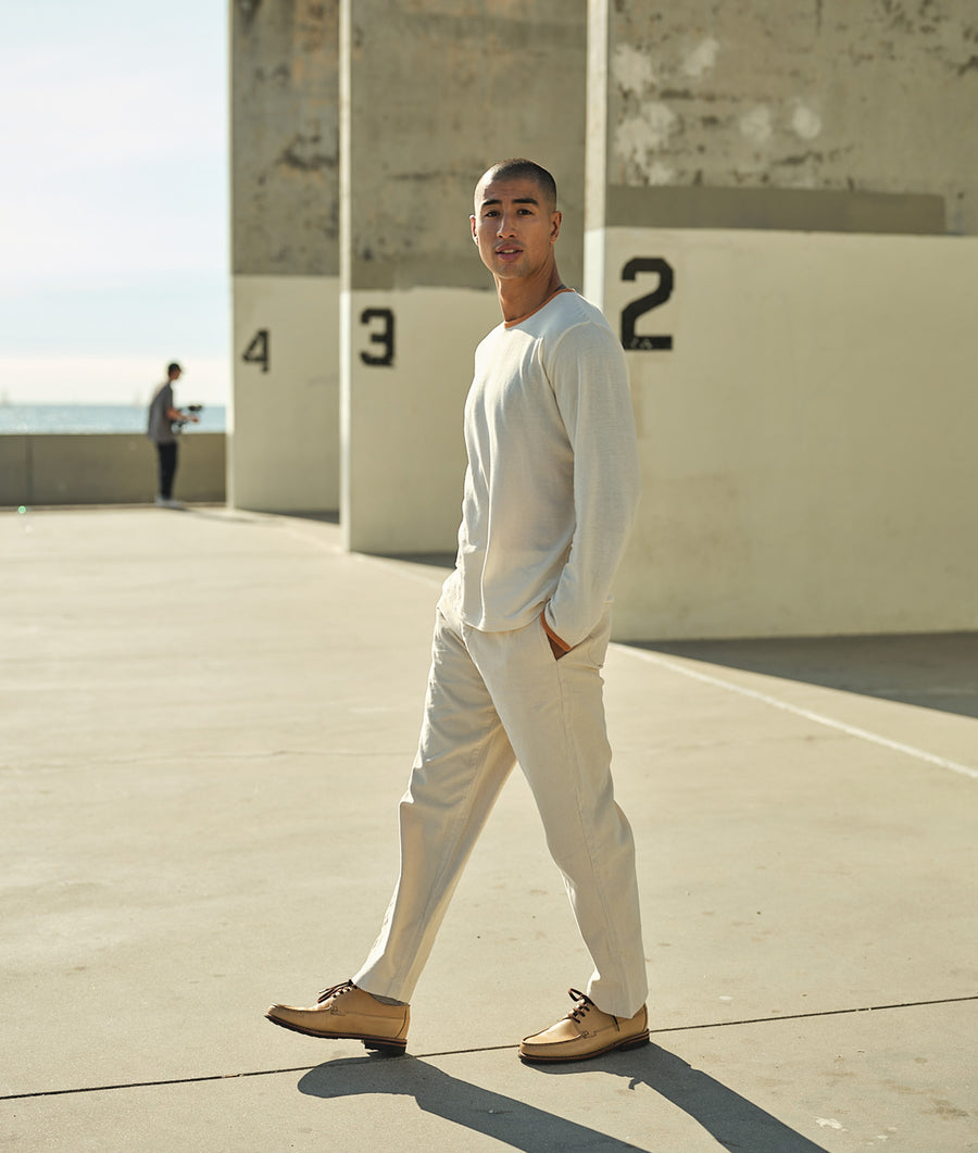 A person wearing a white long-sleeve shirt and light-colored pants, standing in front of a concrete wall with numbers - Industry of All Nations