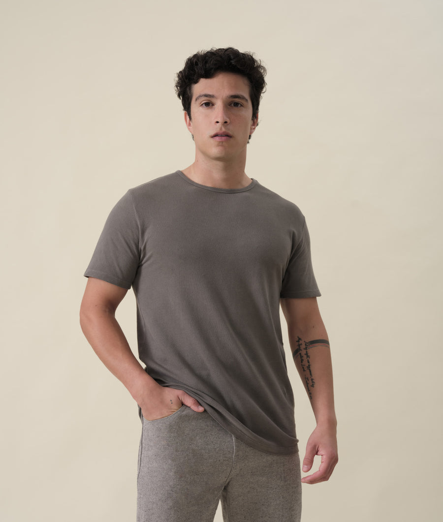 A man wearing a solid gray crew neck t-shirt, standing against a light colored background. - Industry of All Nations