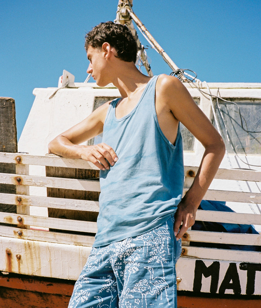 A young person wearing a light blue tank top and patterned pants, standing in front of a wooden structure with 'MAT' written on it - Industry of All Nations.