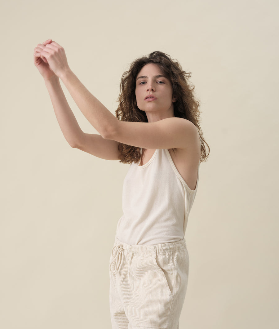 A person with curly brown hair wearing a white sleeveless top, standing against a beige background - Industry of All Nations