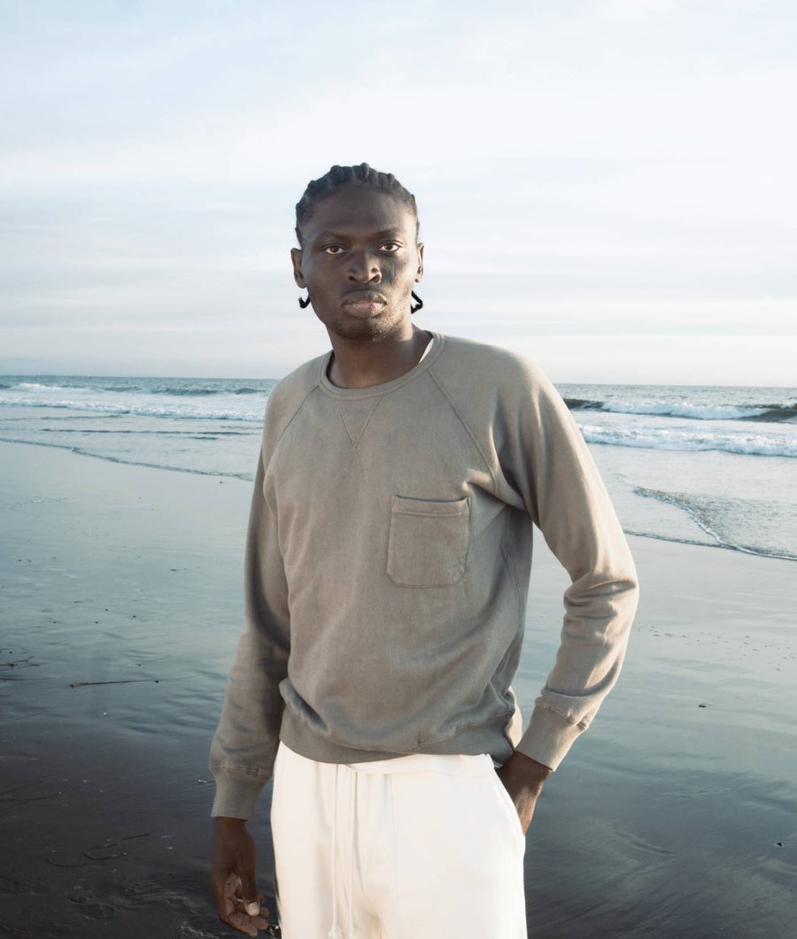 A man wearing a light gray sweatshirt standing on a beach overlooking the ocean - Industry of All Nations
