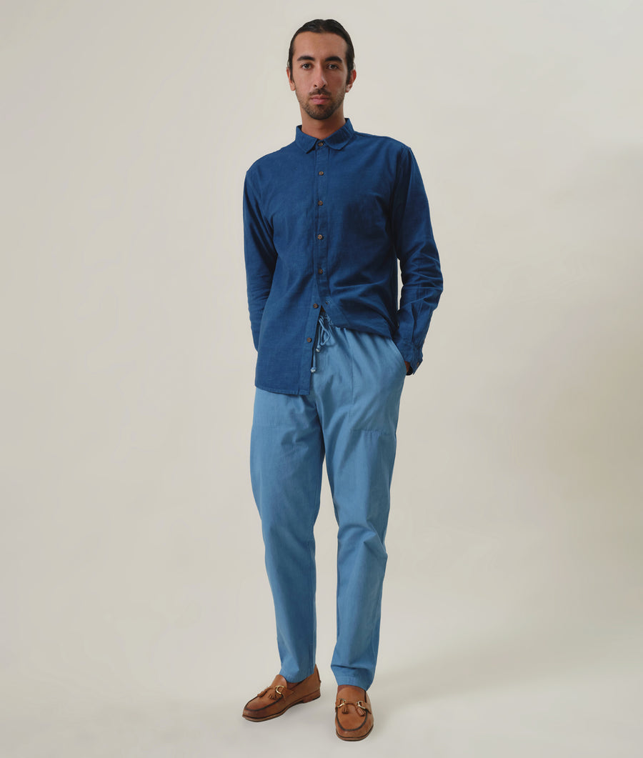 A man wearing a long-sleeved blue shirt and matching blue pants, standing in front of a light background with brown leather shoes - Industry of All Nations