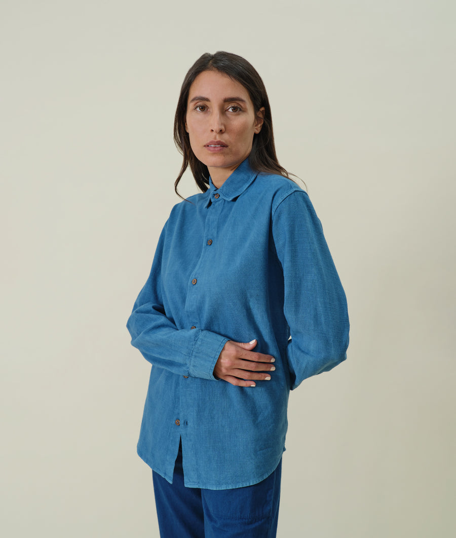 A woman wearing a blue cotton button-up shirt in a frontal pose, with hands crossed - Industry of All Nations