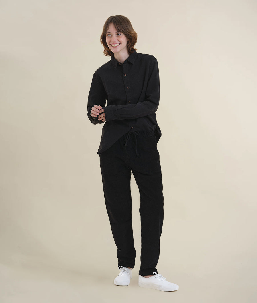 A smiling woman wearing a black organic cotton jumpsuit, standing in a plain setting - Industry of All Nations