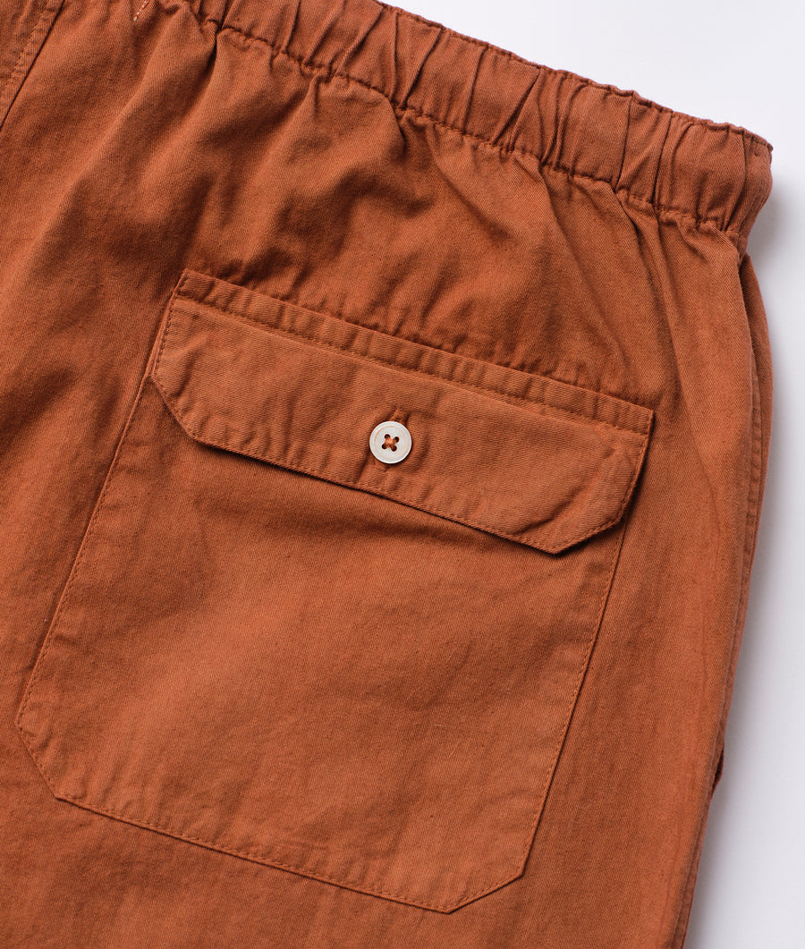 Rust-colored cotton pants with a pocket and button closure, displayed in a detail shot - Industry of All Nations